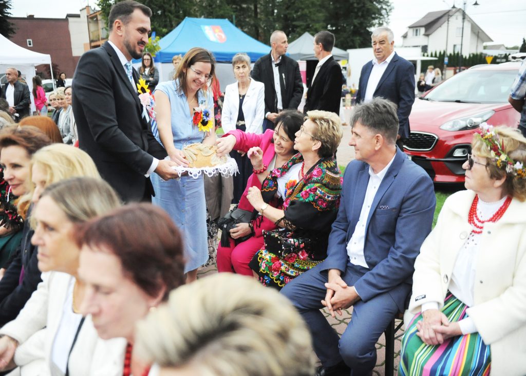 Grupa ludzi na zewnątrz, częstują się chlebem. Kolorowe ubrania, niebieski namiot w tle. Samochód i budynki w tle.