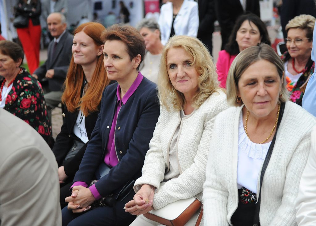 Grupa ludzi siedzi na zewnątrz, większość to kobiety. Ubrania w jasnych i ciemnych kolorach, w tle rozmyte postaci. Kolorowe kwiatowe wzory na ubraniach.