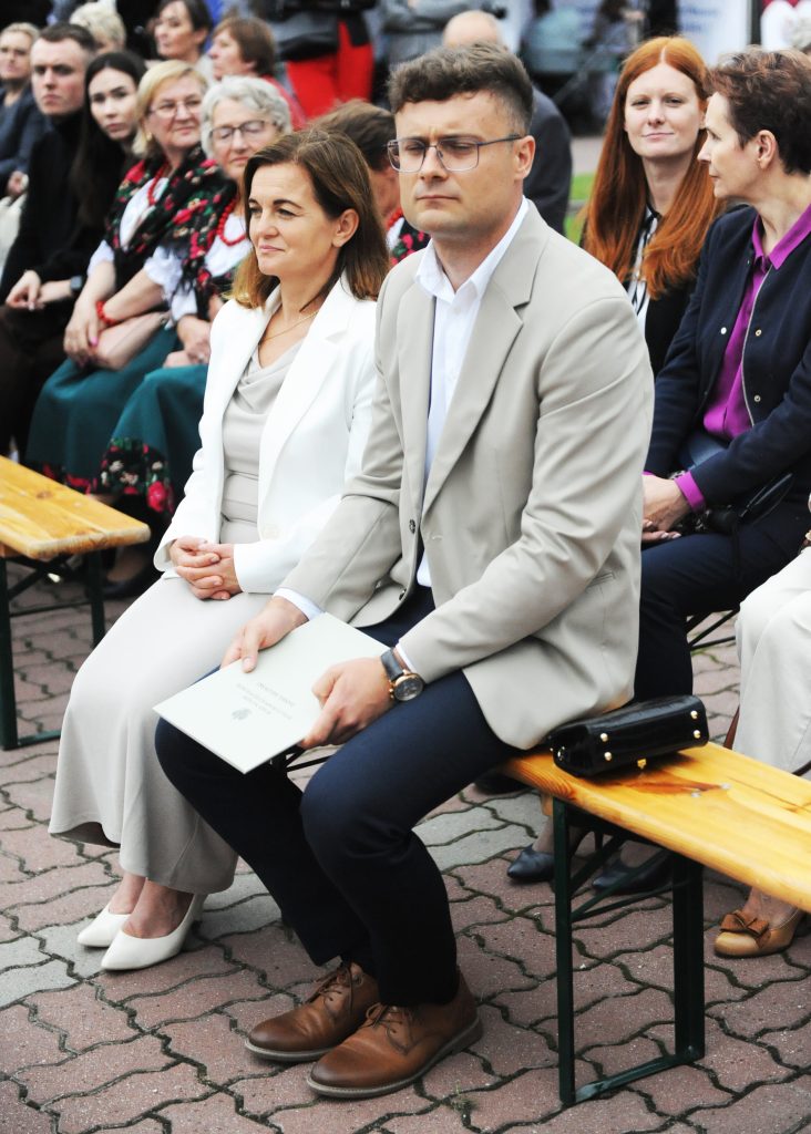 Grupa ludzi siedzi na drewnianych ławkach, na zewnątrz. Dwie osoby na pierwszym planie w jasnych garniturach. Tło wypełnia tłum w kolorowych ubraniach.