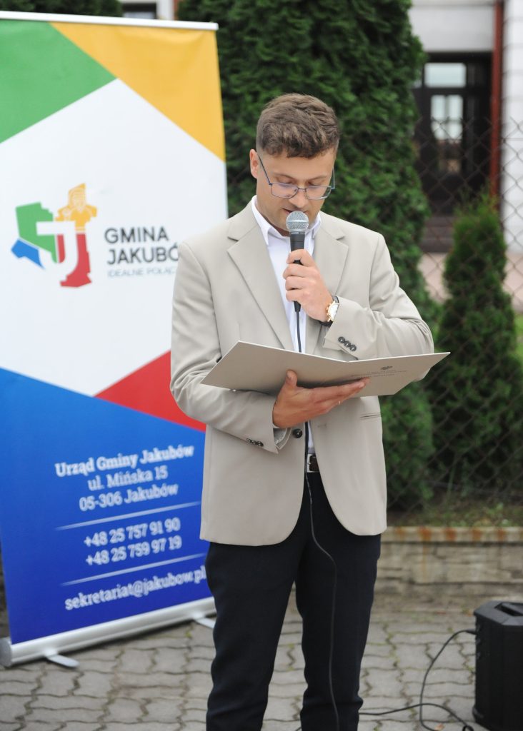 Nieznany mężczyzna w beżowej marynarce mówi do mikrofonu. W tle baner z logo i informacją o Gminie Jakubów. Ceglane tło z zielonymi krzewami.