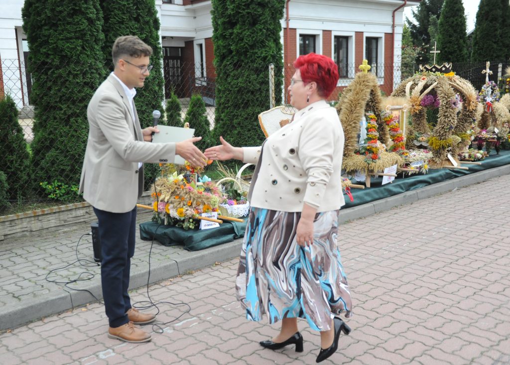 Na zdjęciu kobieta w jasnej marynarce i mężczyzna w beżowej, podają sobie ręce. W tle stoją ozdoby z kłosów i kwiatów. Otoczenie to brukowana ulica i zieleń.