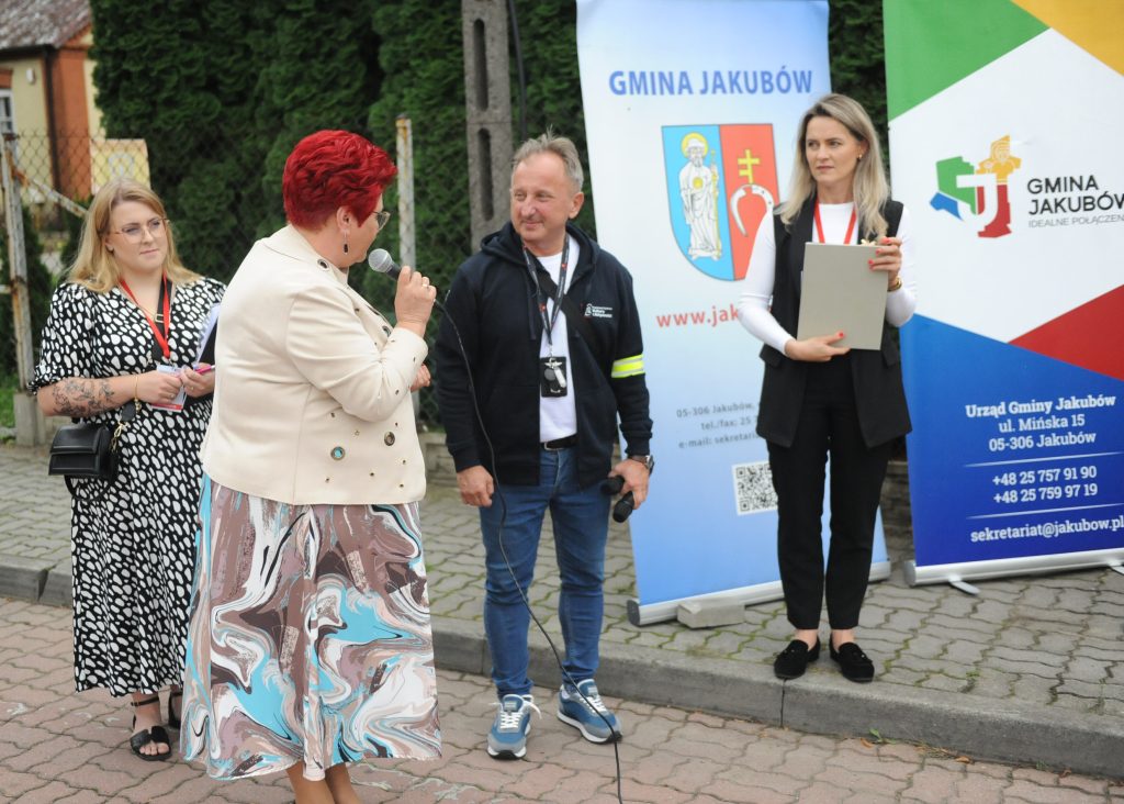 Cztery osoby stoją przed banerem Gmina Jakubów. Kobieta w jasnej kurtce mówi do mikrofonu. Tło to zielone drzewa i baner.