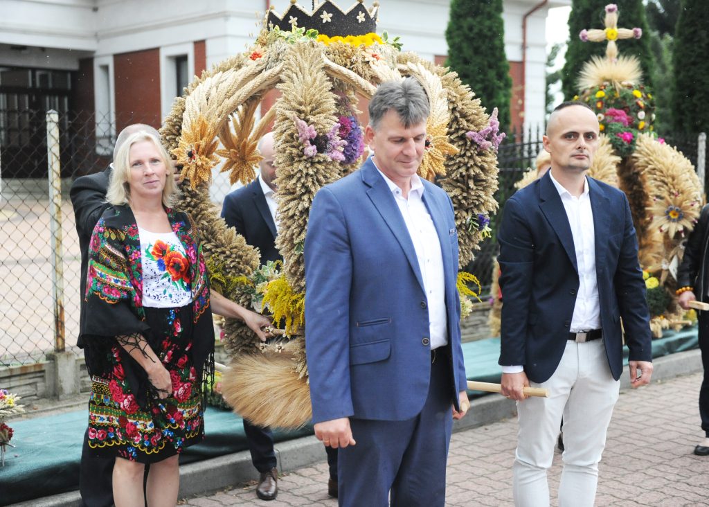 Grupa ludzi w tradycyjnych strojach stoi przed ozdobną koroną dożynkową z kłosów zbóż. Tło stanowi ogrodzenie i budynek. Kolory to głównie złote, zielone i czerwone.