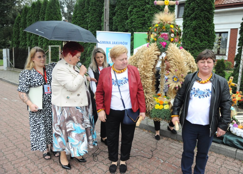 Grupa kobiet stoi przed kolorowym dożynkowym wieńcem ze zbóż i kwiatów. Jedna z nich trzyma mikrofon, inna parasol. W tle drzewa i budynek.