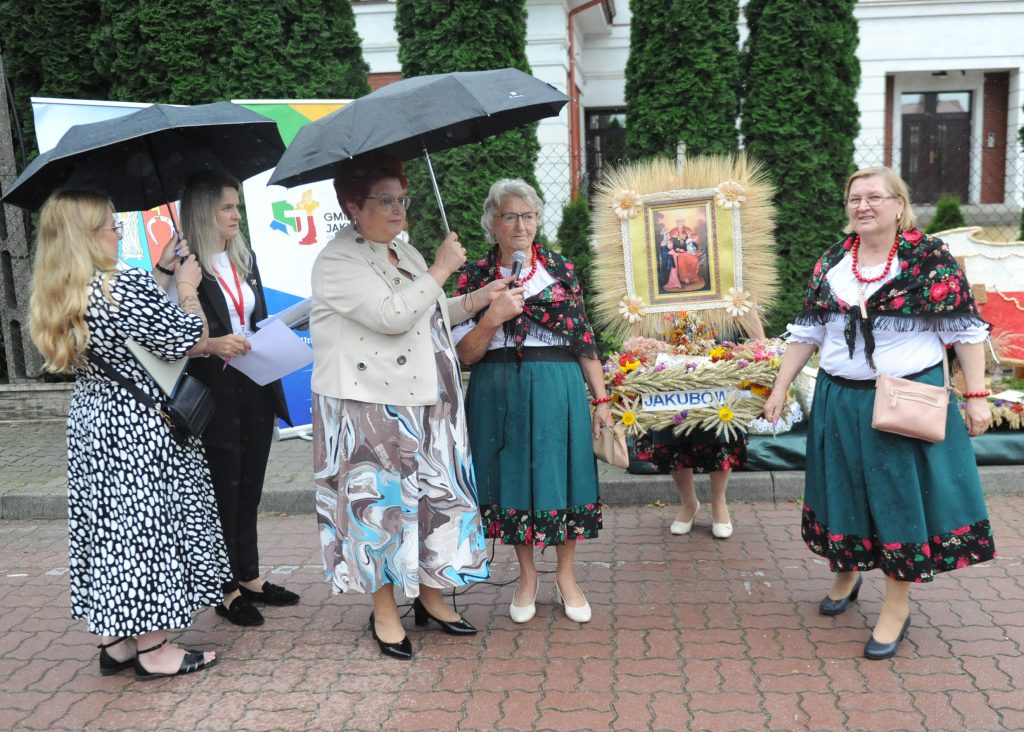 Pięć osób pod parasolami, trzy w tradycyjnych strojach ludowych. Bukiet kwiatów i snop zboża w tle. Tło zielone drzewka i biały budynek.