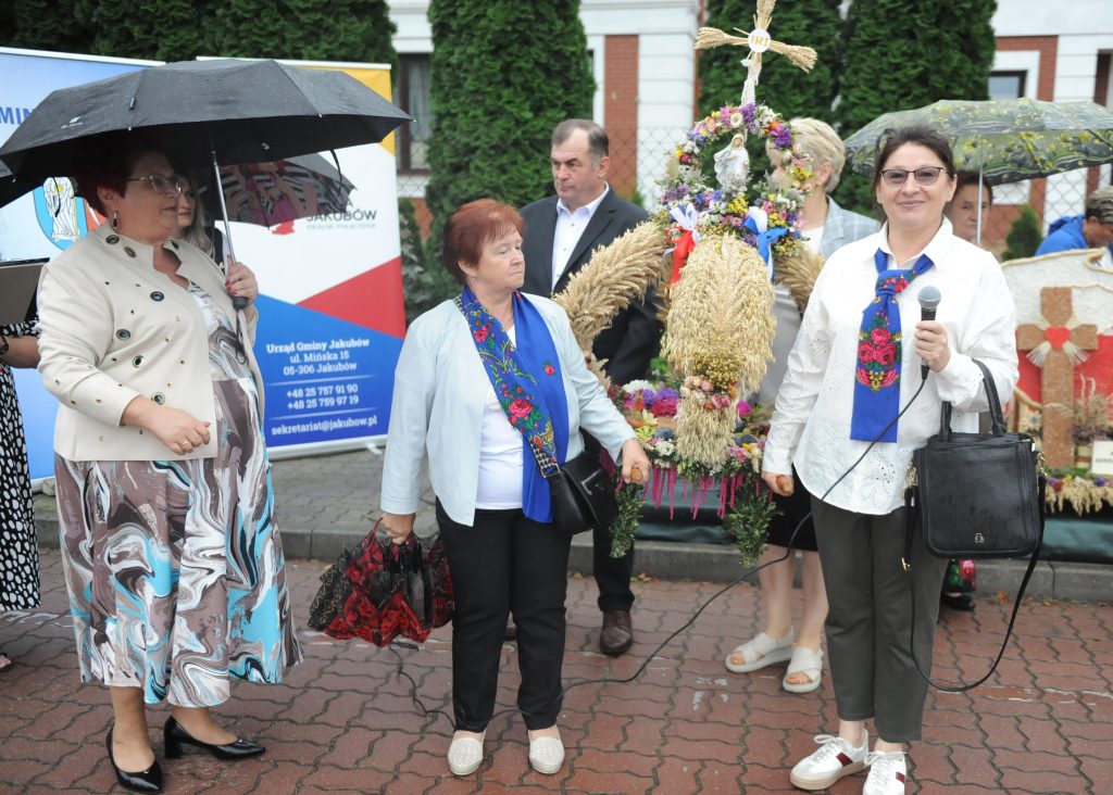 Grupa osób pod parasolami przy kolorowych wieńcach dożynkowych. W tle zielone krzewy i baner. Kobieta z mikrofonem mówi.
