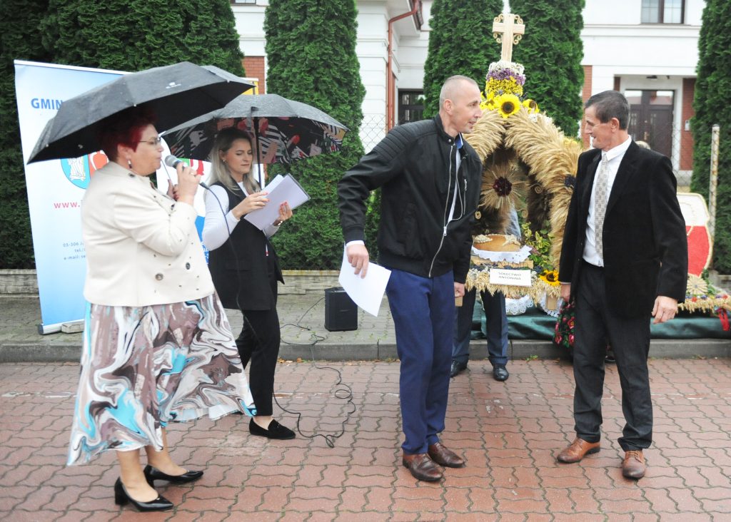 Grupa ludzi pod parasolami na tle ozdobnego wieńca i zieleni. Kobieta przemawia do mikrofonu. Na ziemi czerwona kostka brukowa.