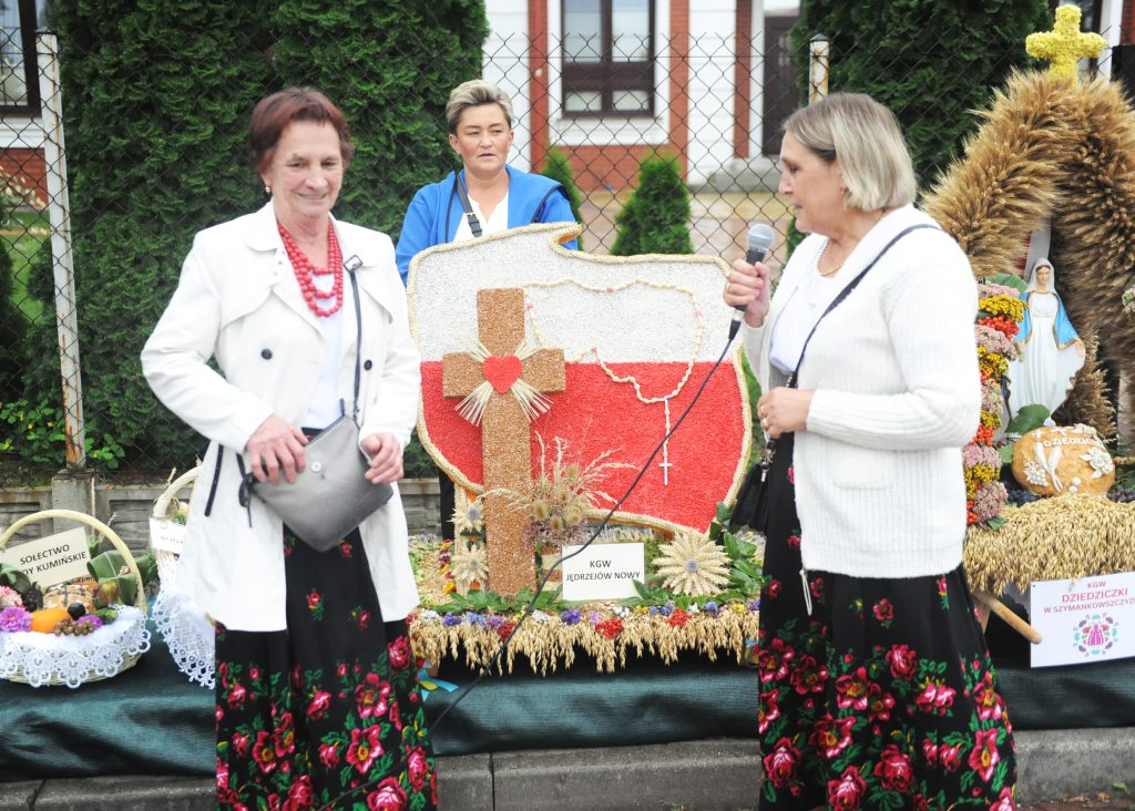 Trzy kobiety w tradycyjnych strojach stoją przed dekoracją z mapą Polski i krzyżem. W tle zielone krzewy.