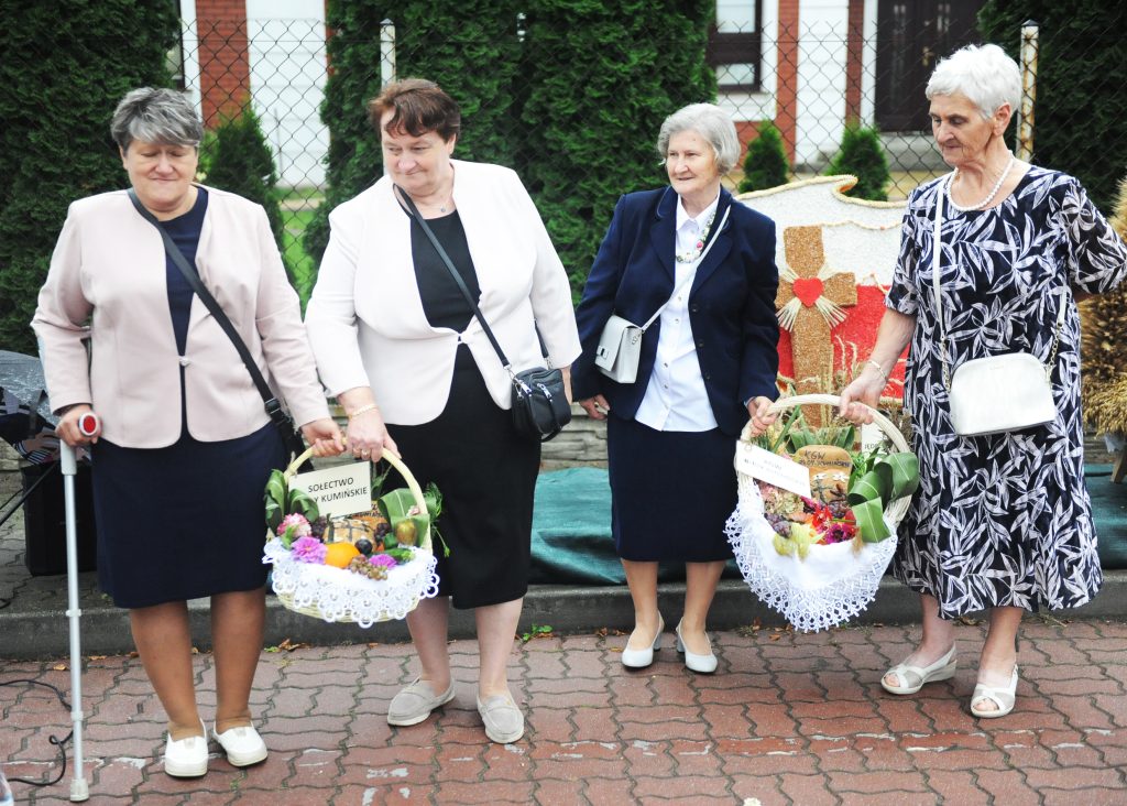 Cztery kobiety w eleganckich ubraniach trzymają ozdobne kosze z darami. Tło: zielone krzewy, metalowy płot. Kobiety mają białe i granatowe stroje.