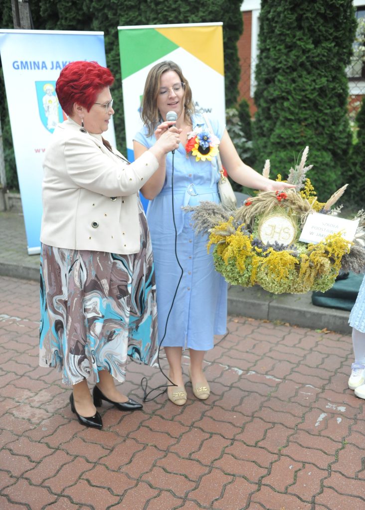 Dwie kobiety podczas ceremonii, jedna trzyma mikrofon, druga kosz z kwiatami. Kobiety są na chodniku, za nimi zielone krzewy i banery. Ubrania w jasnych kolorach, ustawienie oficjalne.