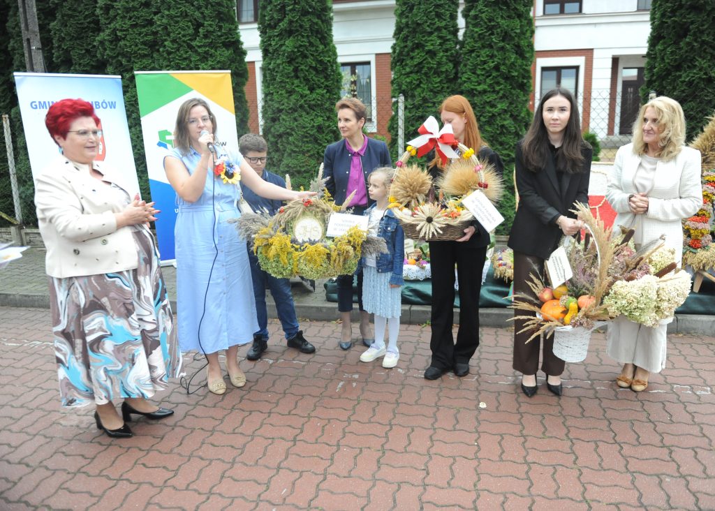 Grupa osób z koszami pełnymi zbóż i kwiatów, stoją na tle zielonych krzewów. Osoba w niebieskiej sukience przemawia do mikrofonu. Obok znajdują się kolorowe banery.