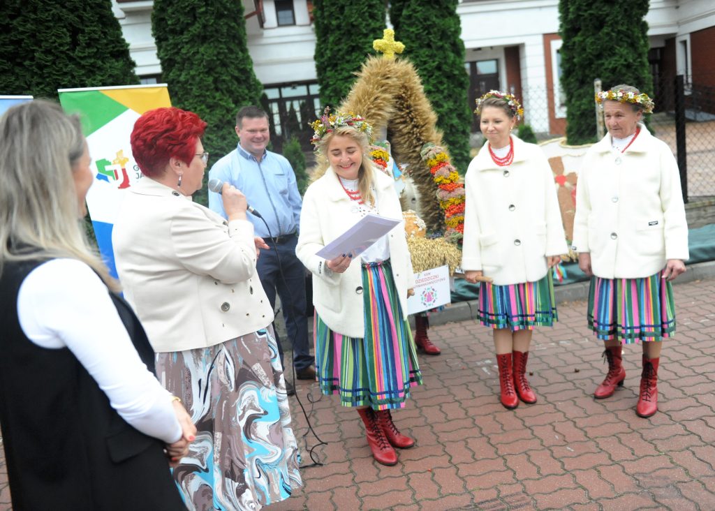 Grupa kobiet w kolorowych strojach ludowych z wiankami na głowach. W tle zielone iglaki i biały budynek. Jedna z kobiet mówi do mikrofonu.