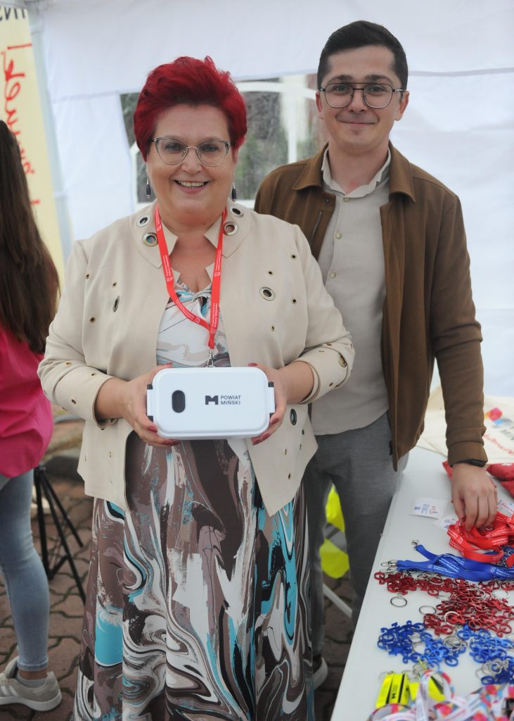 Na zdjęciu starsza kobieta w beżowej garderobie trzyma biały gadżet z napisem Powiat Miński. Obok niej stoi mężczyzna w brązowej kurtce. W tle biały namiot i stół z kolorowymi breloczkami.