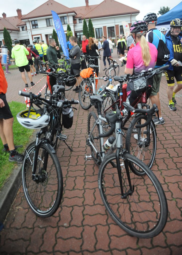 Grupa rowerzystów na kostce brukowej, z rowerami stojącymi w szeregu. Ludzie ubrani w kolorowe stroje sportowe. W tle budynek z czerwoną dachówką i zielone krzewy.