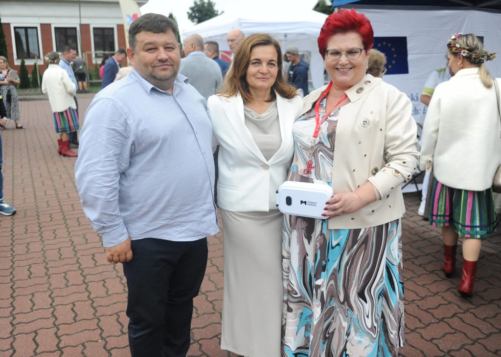Na zdjęciu troje uśmiechniętych osób na tle ludzi. Kobieta z lewej ma czerwone włosy, trzyma pudełko. Płyta chodnikowa, budynki w tle.