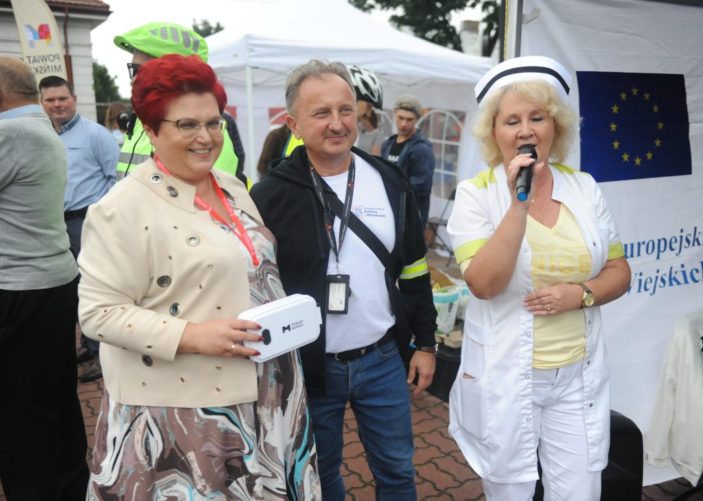 Trzy osoby pozują w plenerze obok białego namiotu. Kobieta z mikrofonem jest ubrana na biało. W tle widoczne logo UE.