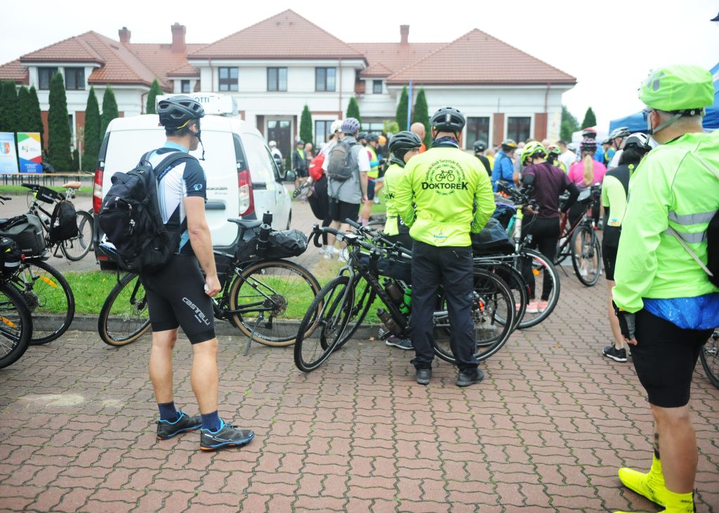 Grupa rowerzystów w kaskach stoi na placu przed białym budynkiem. Mają kolorowe kurtki, głównie w odcieniach zieleni. W tle samochód dostawczy.
