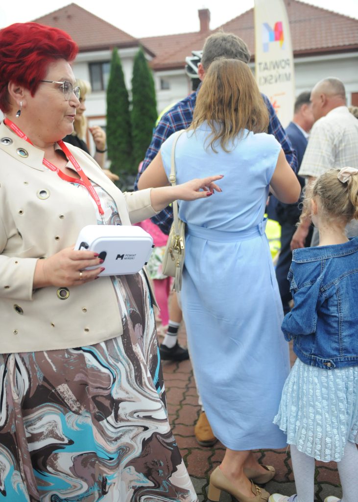 Kobieta z czerwoną fryzurą i jasnym płaszczem stoi w tłumie, trzymając białą torbę z napisem. Obok niej kobieta w niebieskiej sukience, dziecko w niebieskiej kurtce. W tle budynki z czerwonym dachem.