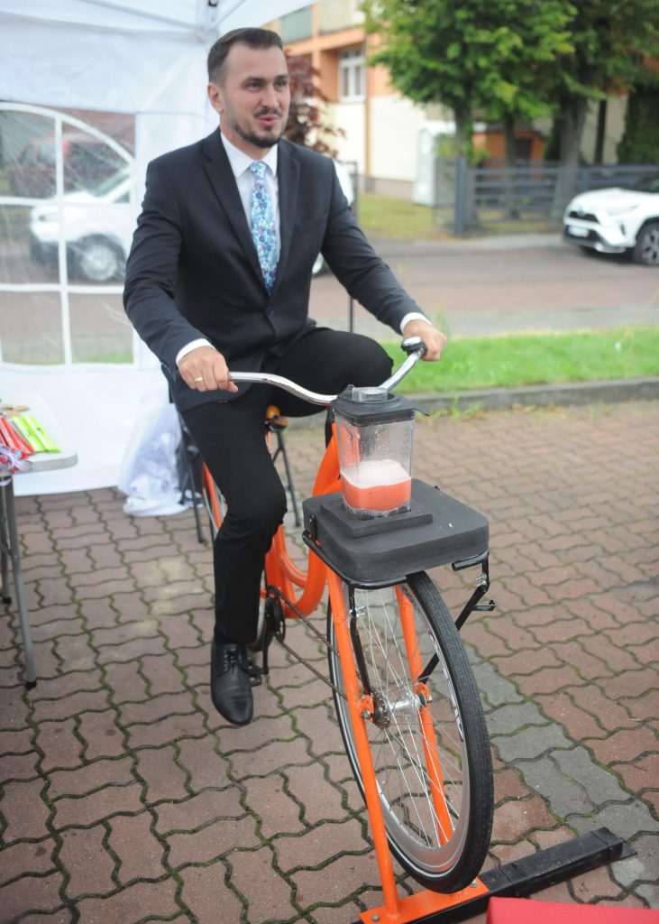 Mężczyzna w garniturze na pomarańczowym rowerze z blenderem. Tło to zieleń i zaparkowane auta. Na białym stole kolorowe akcesoria.