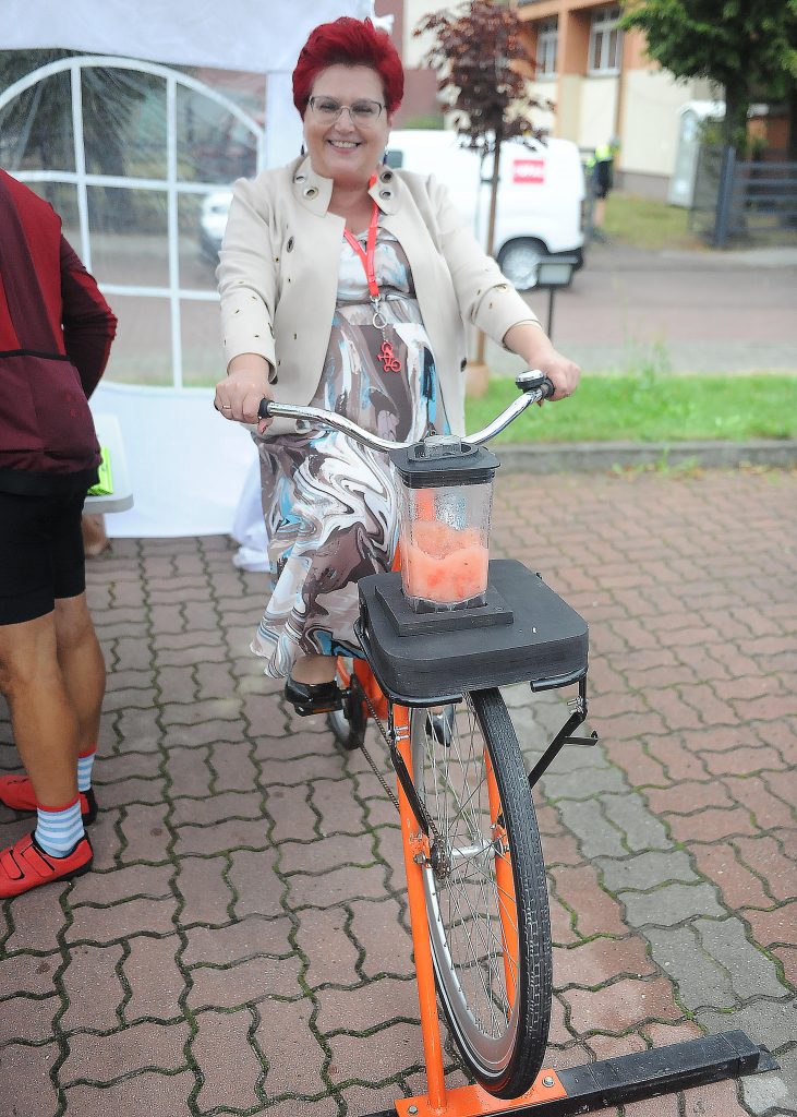 Osoba na rowerze z blenderem przygotowuje smoothie. Ma na sobie kolorową sukienkę i jasną marynarkę. Tło to ulica z drzewami.