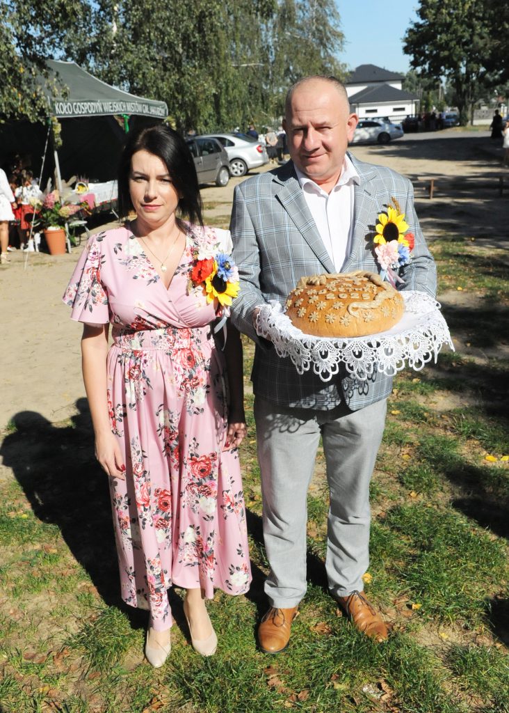 Para stoi na zewnątrz, mężczyzna trzyma chleb na serwetce. Kobieta w różowej sukience, mężczyzna w marynarce. W tle namiot i drzewa.