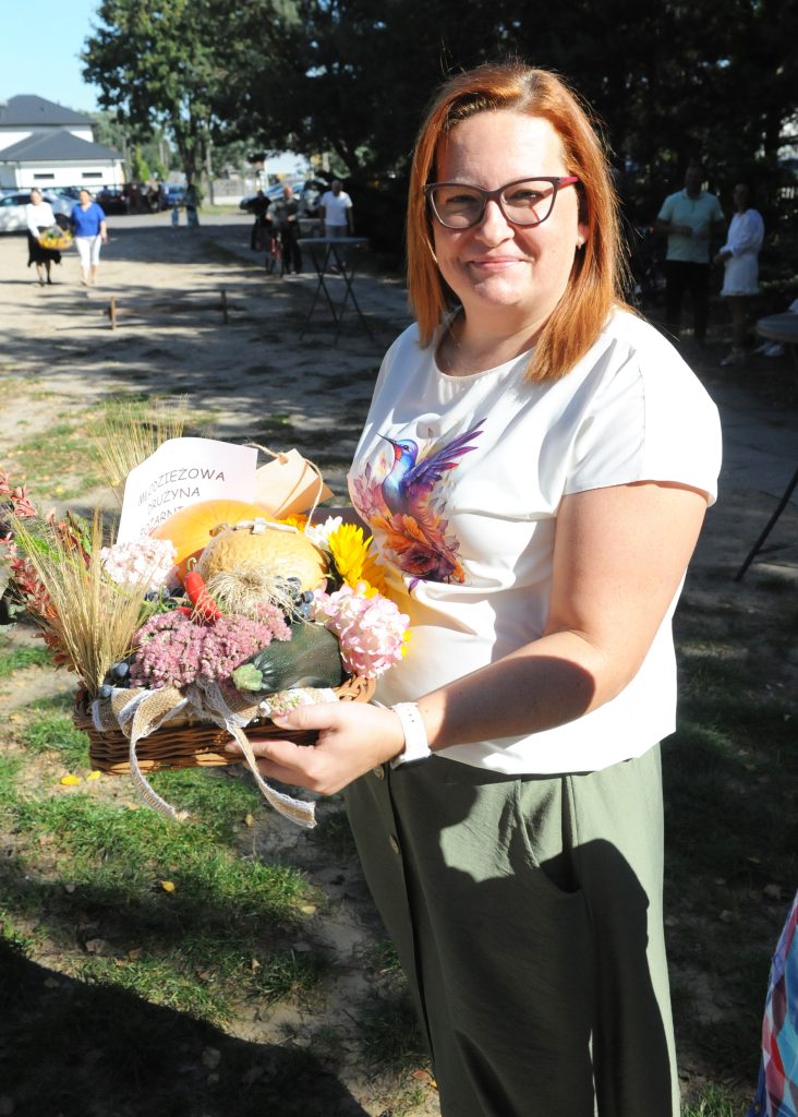 Kobieta w okularach trzyma ozdobny koszyk z kwiatami. Ma białą bluzkę z ptakiem, stoi na zewnątrz przy słonecznej pogodzie. W tle drzewa i ludzie.