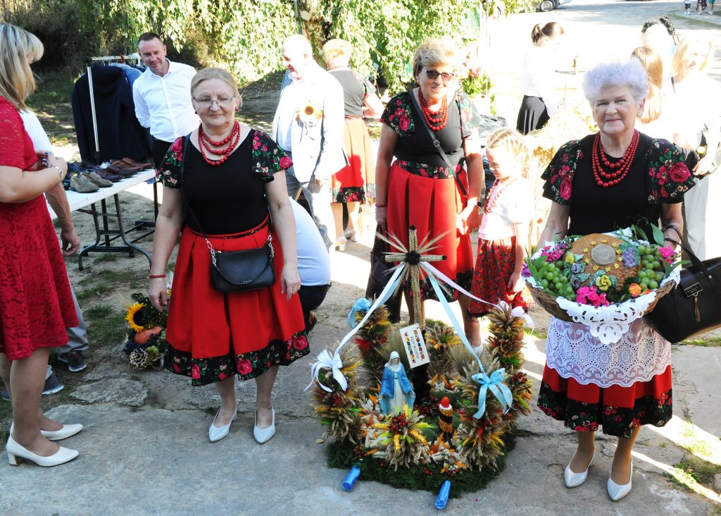 Grupa osób w tradycyjnych strojach ludowych z czerwonymi spódnicami stoi na tle zieleni. Trzymają ozdobne wieńce z kwiatów i owoców. Wokół nich znajdują się inni uczestnicy wydarzenia.