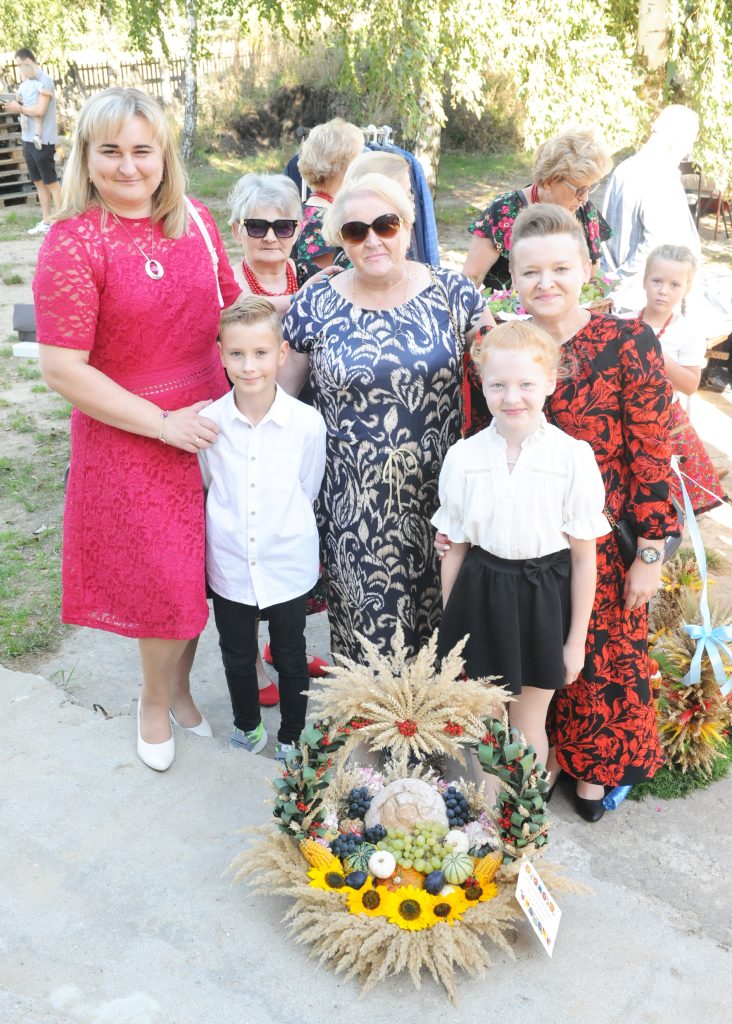 Grupa ludzi pozuje z kolorowym wieńcem dożynkowym z owocami i zbożem. Dominują wzorzyste i czerwone stroje. Tło to zielona przestrzeń na świeżym powietrzu.