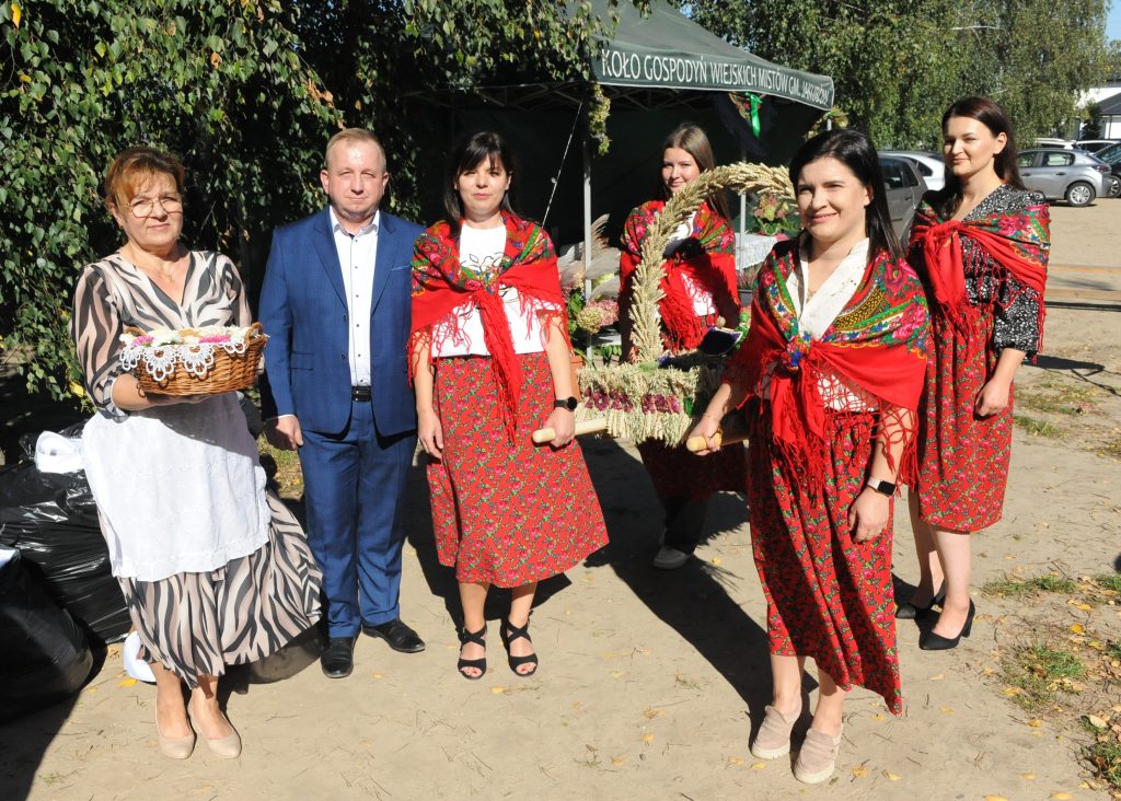 Grupa osób w stroju ludowym, z czerwonymi chustami, stoi na zewnątrz. Jedna osoba trzyma kosz, w tle zielone drzewa i namiot.