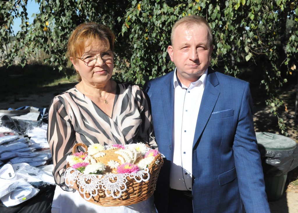 Na zdjęciu są dwie osoby stojące obok siebie. Kobieta trzyma wiklinowy kosz z kwiatami na tle zielonych drzew. Mężczyzna jest w niebieskim garniturze.