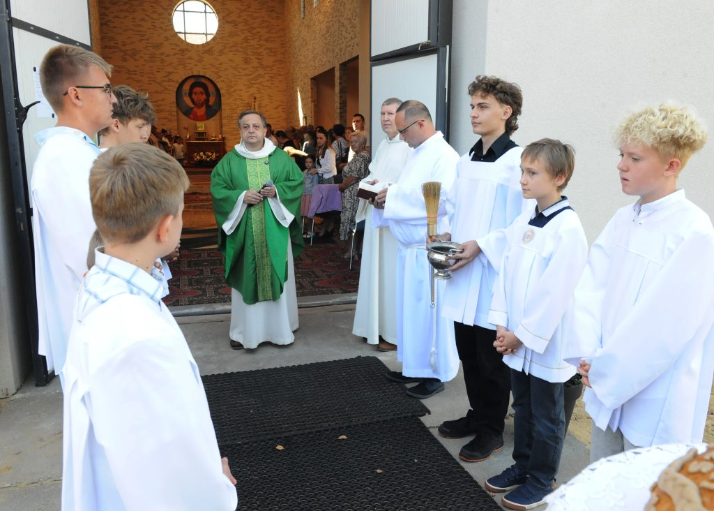 Na zdjęciu widoczna jest grupa ministrantów oraz kapłan w zielonych szatach liturgicznych. Mężczyźni stoją przy wejściu do kościoła z ceglastymi ścianami. Tło stanowi wnętrze kościoła z okrągłym witrażem