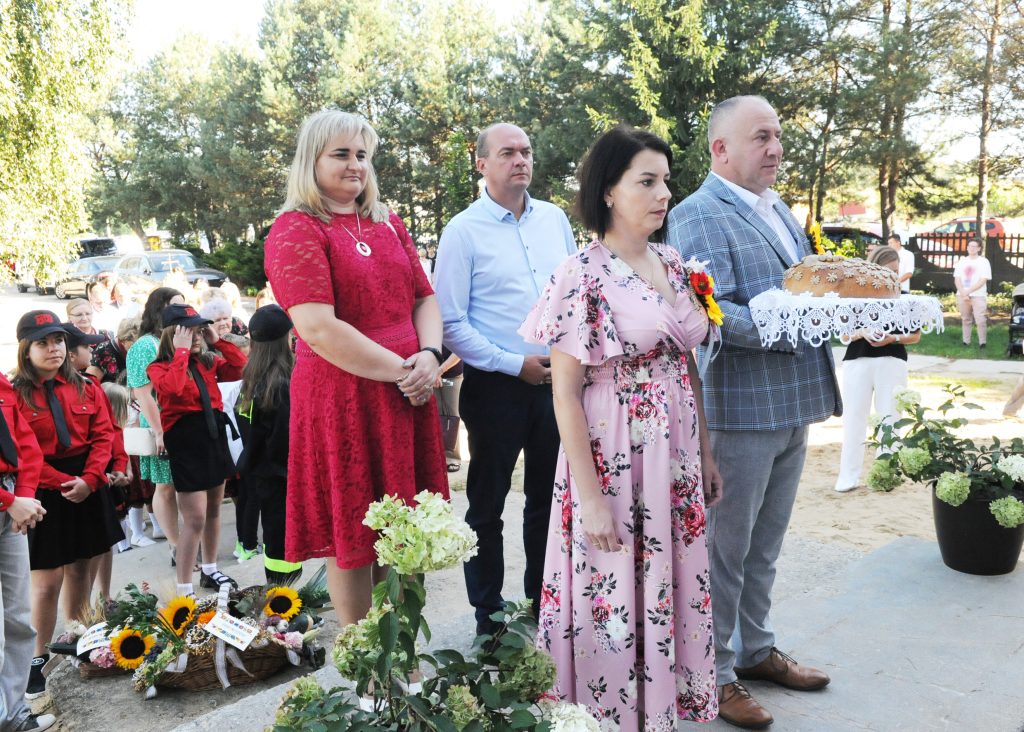 Grupa ludzi na zewnątrz, trzymająca chleb na ozdobionej serwetce. Tło z drzewami i zebranymi ludźmi. Jasne stroje i kwiaty.