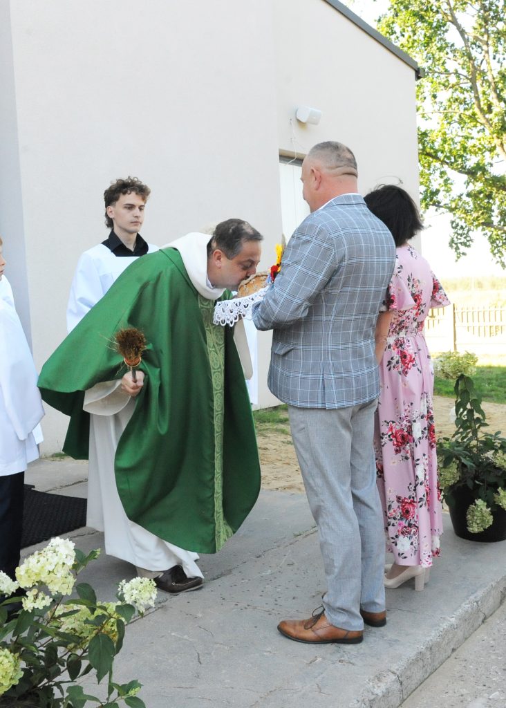 Mężczyzna w zielonej szacie całuje chleb, trzymając kropidło. Obok stoją kobieta w kwiecistej sukni i mężczyzna w szarej marynarce. W tle widać budynek i drzewa.