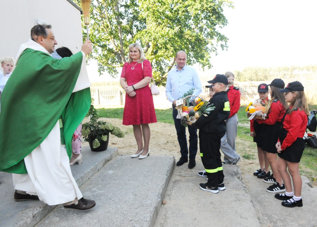 Ksiądz w zielonej stule błogosławi dzieci w strojach strażackich. Obok kobieta w czerwonej sukience. W tle zielone drzewo.