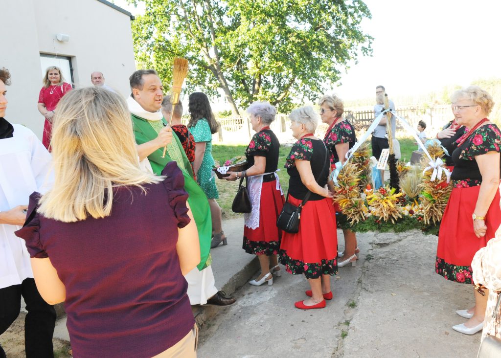 Grupa osób stoi na dworze, ksiądz w zielonym stroju błogosławi wieńce dożynkowe. Kobiety w ludowych strojach z czerwonymi spódnicami. Tło stanowią drzewa i biały budynek.