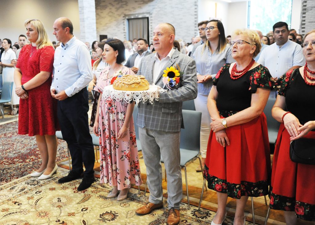 Grupa ludzi stoi w sali, jeden z nich trzyma chleb na serwecie. W tle widać jasne ściany i okno. Ludzie są ubrani w różnorodne stroje, niektórzy w tradycyjne.