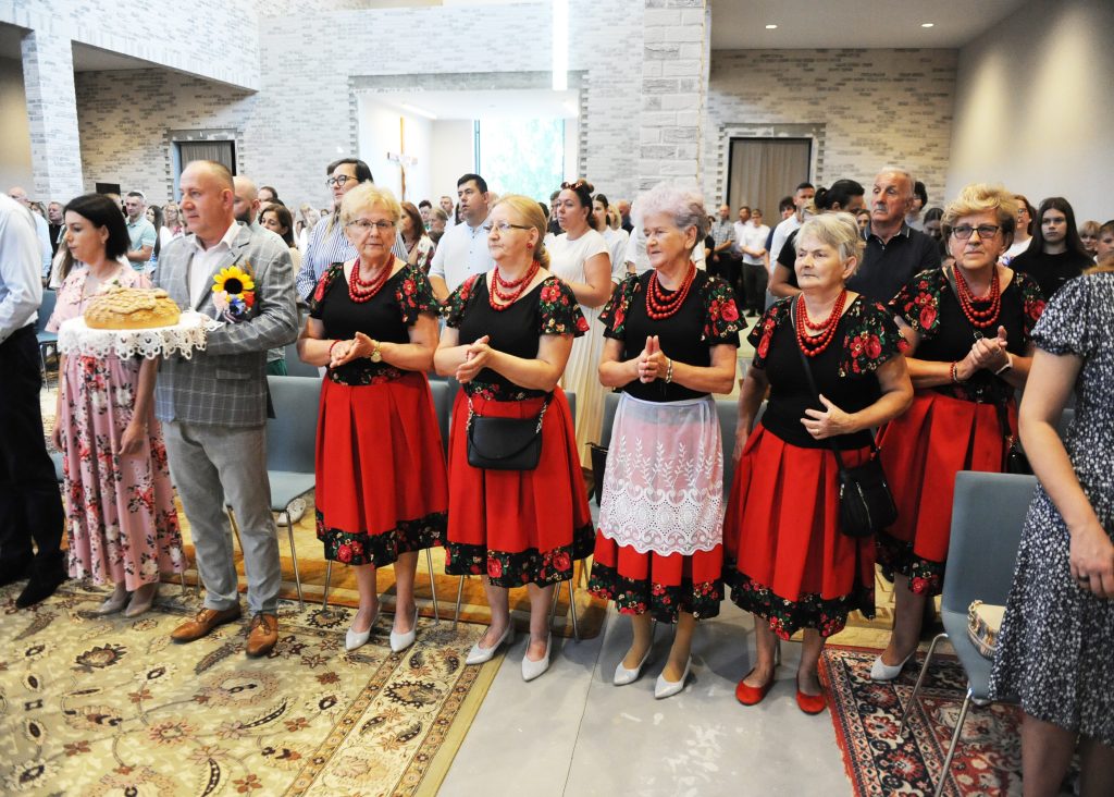 Grupa osób ubranych w tradycyjne stroje ludowe stoi we wnętrzu budynku o jasnych ścianach. Kobiety mają czerwone spódnice i czarne bluzki, mężczyzna niesie chleb na tacy. W tle widać zebrań publiczność i krzyż.