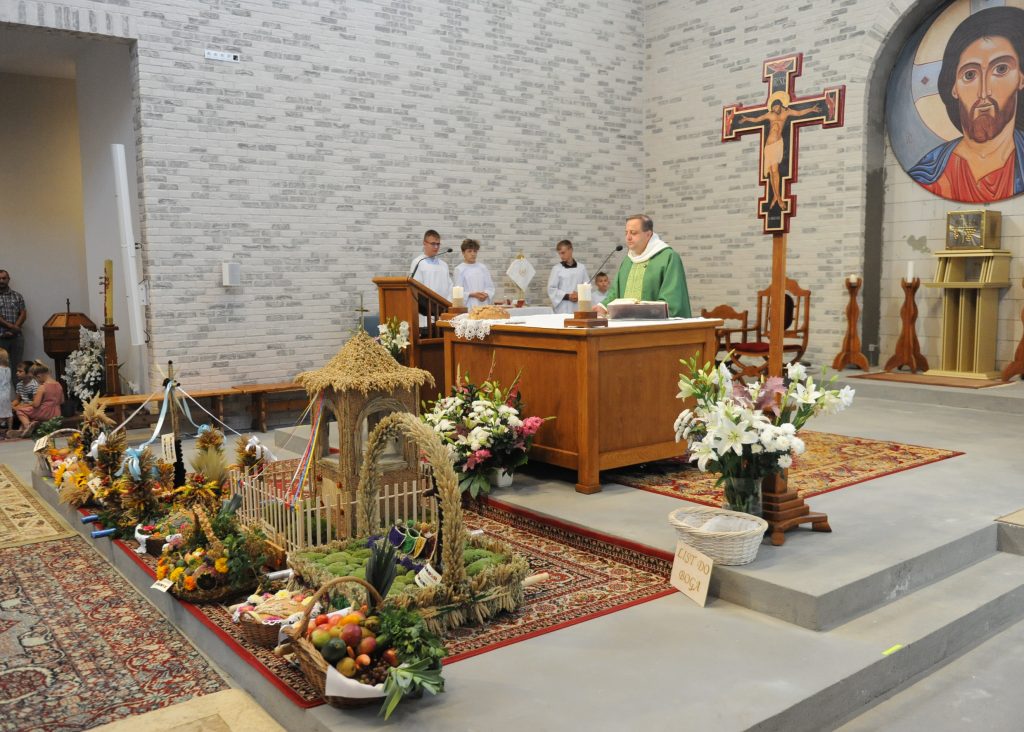 Ksiądz w zielonej szacie odprawia mszę przy ołtarzu. Obok dekoracje z wieńcami, kosze z owocami i kwiaty. Tło stanowi jasna ściana z ikoną.