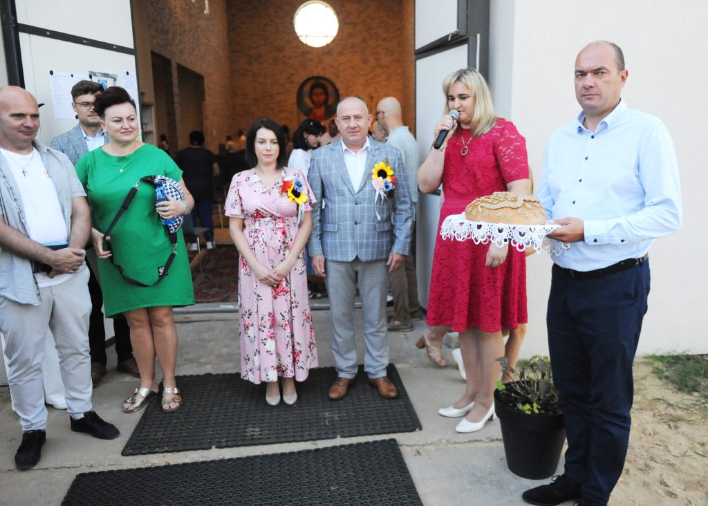 Grupa ludzi przed budynkiem, trzyma mikrofon i chleb. Kobiety w kolorowych sukienkach, mężczyzna w szarym garniturze. W tle ceglana ściana z okrągłym oknem.