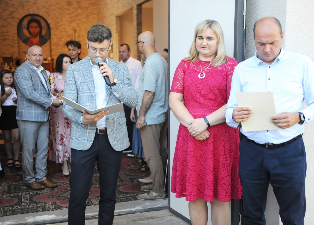 Grupa ludzi stoi w jasnym pomieszczeniu przy otwartych drzwiach. Mężczyzna w marynarce mówi do mikrofonu. Tło zdobi kolorowy wizerunek.