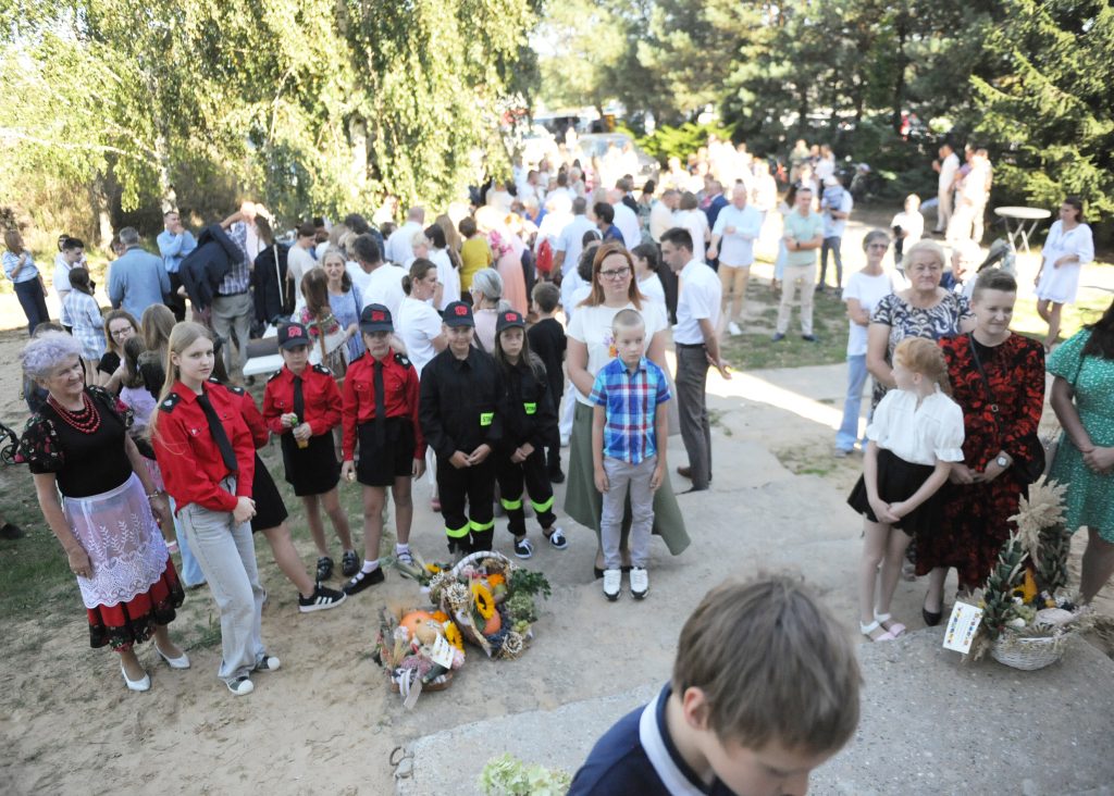 Grupa ludzi zgromadzona na zewnątrz w słoneczny dzień, ubrania w jasnych i kolorowych barwach. Na pierwszym planie młodzież w czerwonych uniformach i koszule w kratę. W tle drzewa i trawa, tworzące naturalne otoczenie.