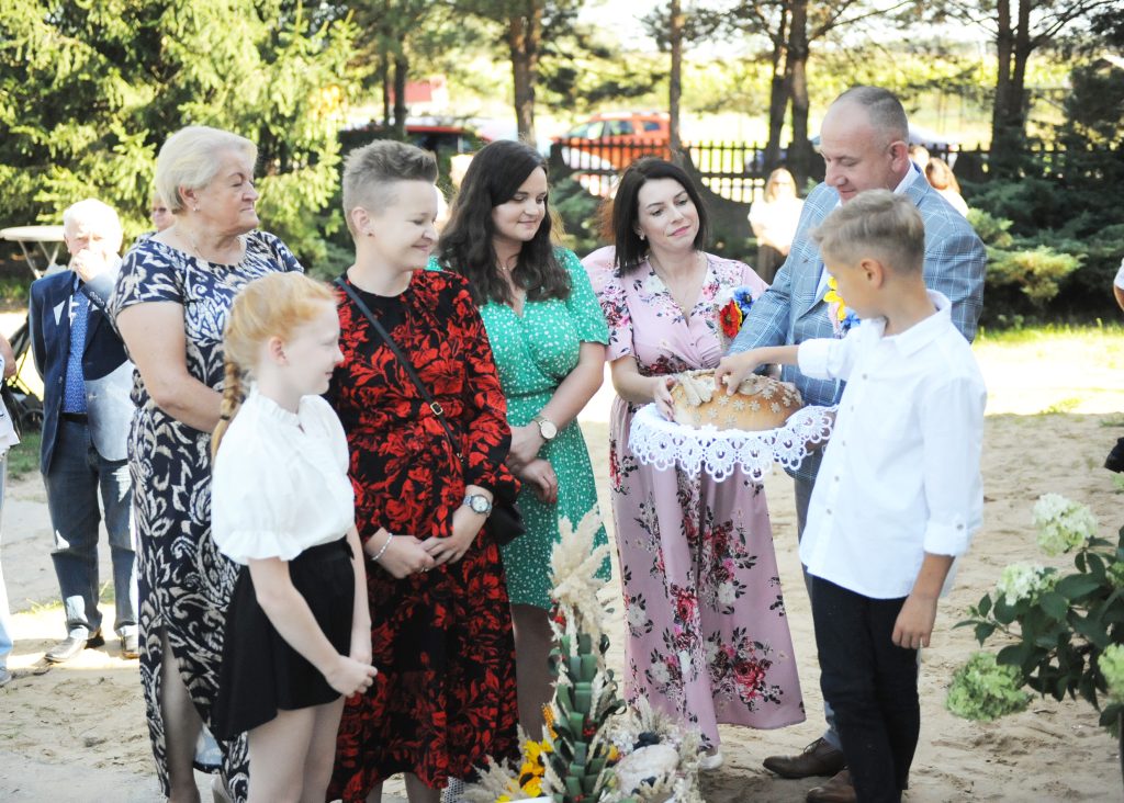 Grupa osób na zewnątrz, zielone tło z drzewami. Dziecko w białej koszuli trzyma talerz z chlebem. Uroczystość na świeżym powietrzu.