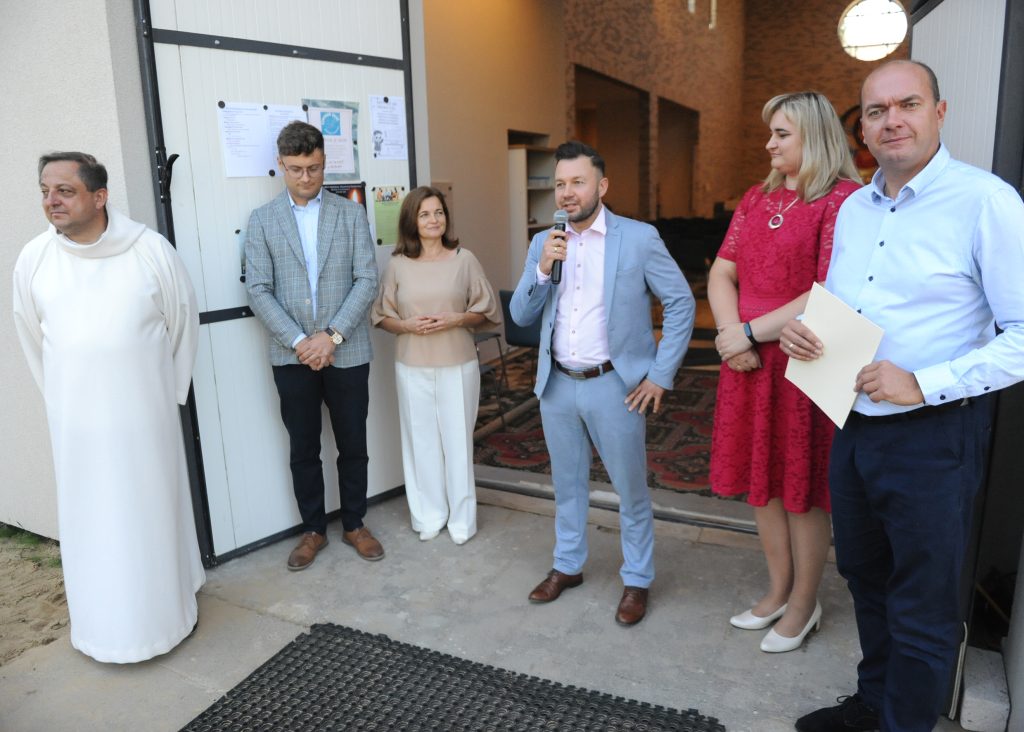 Grupa ludzi stoi przed wejściem do budynku. Mężczyzna z mikrofonem w niebieskim garniturze przemawia. Ściany są jasne, dywan w ciemne wzory.