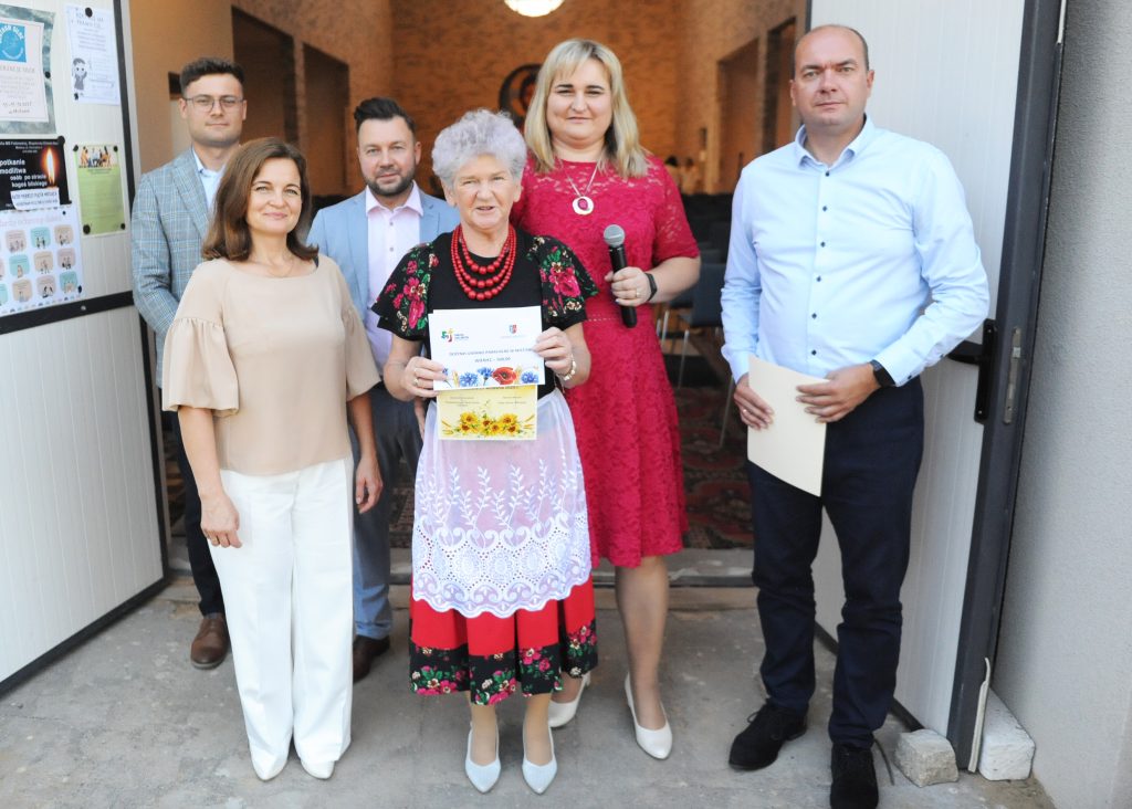 Grupa ludzi stoi przed otwartymi drzwiami, jedno trzyma mikrofon. Osoba w centrum w ludowym stroju z kartką w rękach. Tło jasne, z plakatami na ścianie.