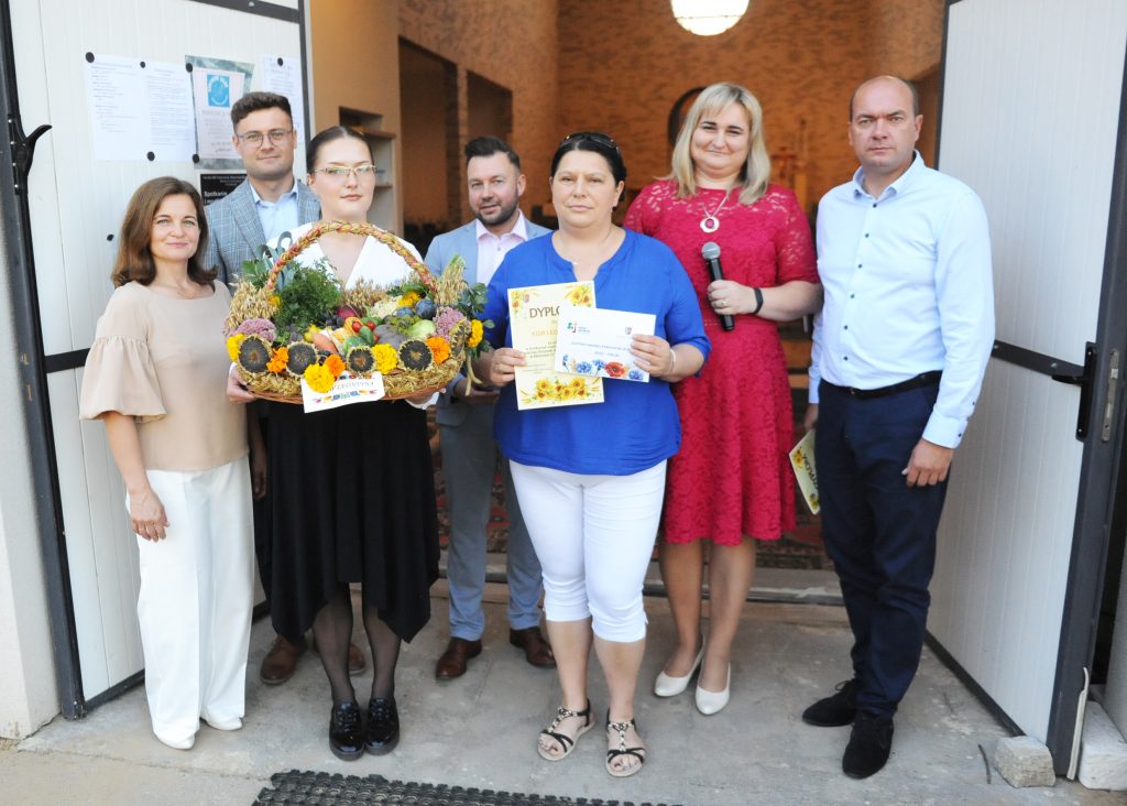 Grupa ludzi stoi przed otwartymi drzwiami budynku. Kobieta trzyma kosz z owocami i warzywami. W tle ceglana ściana.