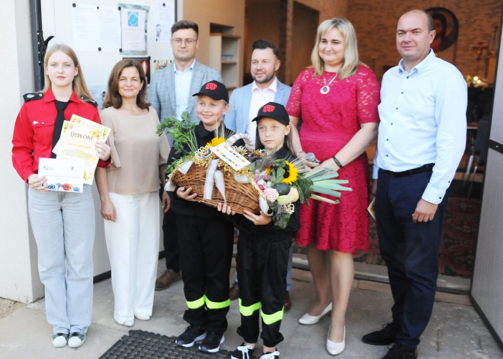 Grupa ludzi pozuje z dyplomem i koszem kwiatów. Ubrania są kolorowe, dominuje czerwień i biel. Tło stanowi wejście budynku.