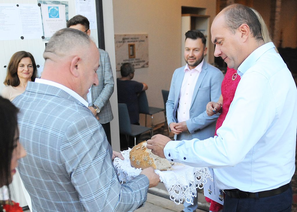 Grupa ludzi stoi, dwaj mężczyźni po lewej trzymają chleb na koronkowej serwetce. Mężczyzna po prawej odłamuje kawałek chleba. W tle osoby siedzą i stoją w jasnym pomieszczeniu.