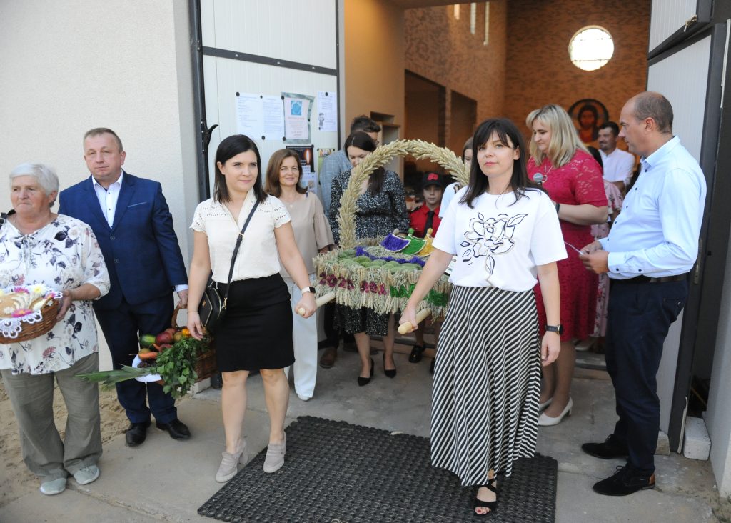 Grupa ludzi wychodzi z budynku, niosąc kolorowe kosze i dekoracje. W centrum kobiety trzymają ozdobioną koronę żniwną. Dominują jasne kolory i elementy kwiatowe na tle jasnych ścian.