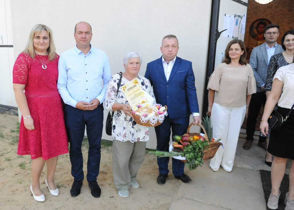 Na zdjęciu grupa osób, środkowa osoba trzyma kosz z owocami i dyplom. Tło to jasna ściana budynku. Ubrania w różnych kolorach, jedna z pań ma czerwoną sukienkę.