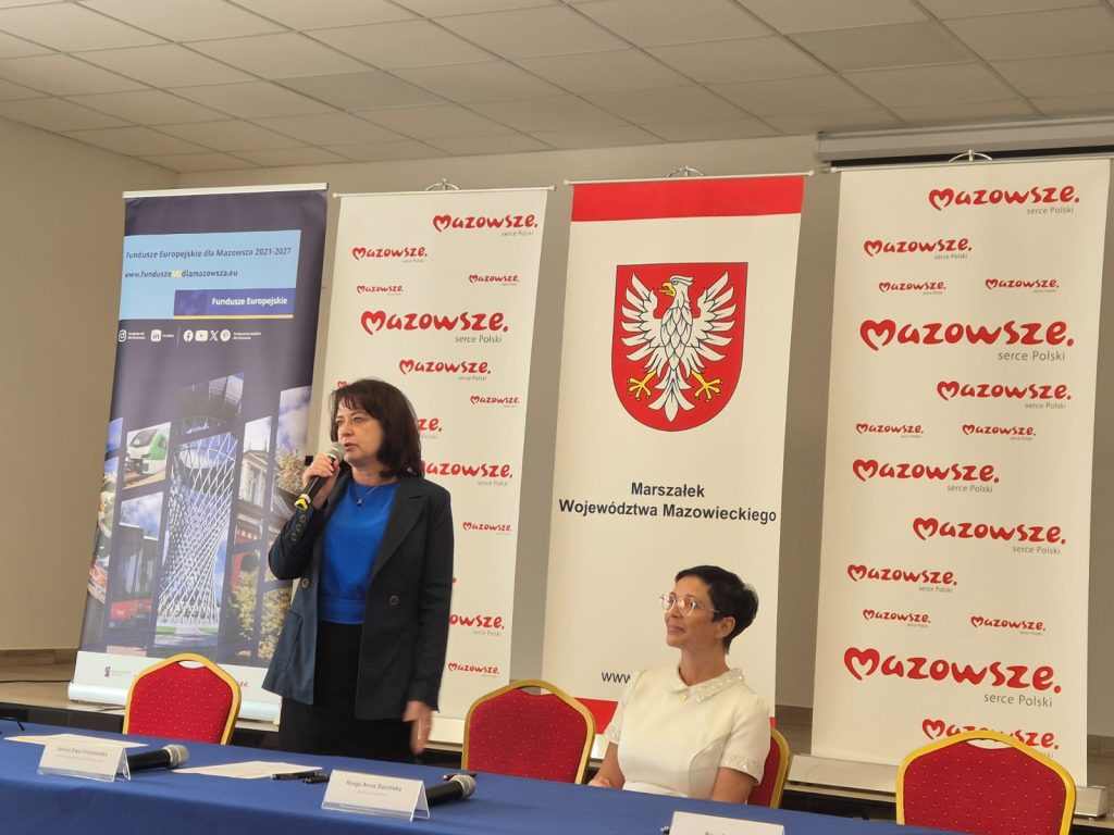 Na zdjęciu kobieta w granatowym ubraniu przemawia przy mikrofonie. Obok siedzi inna kobieta w białej bluzce. W tle widoczne są banery z napisem Mazowsze.
